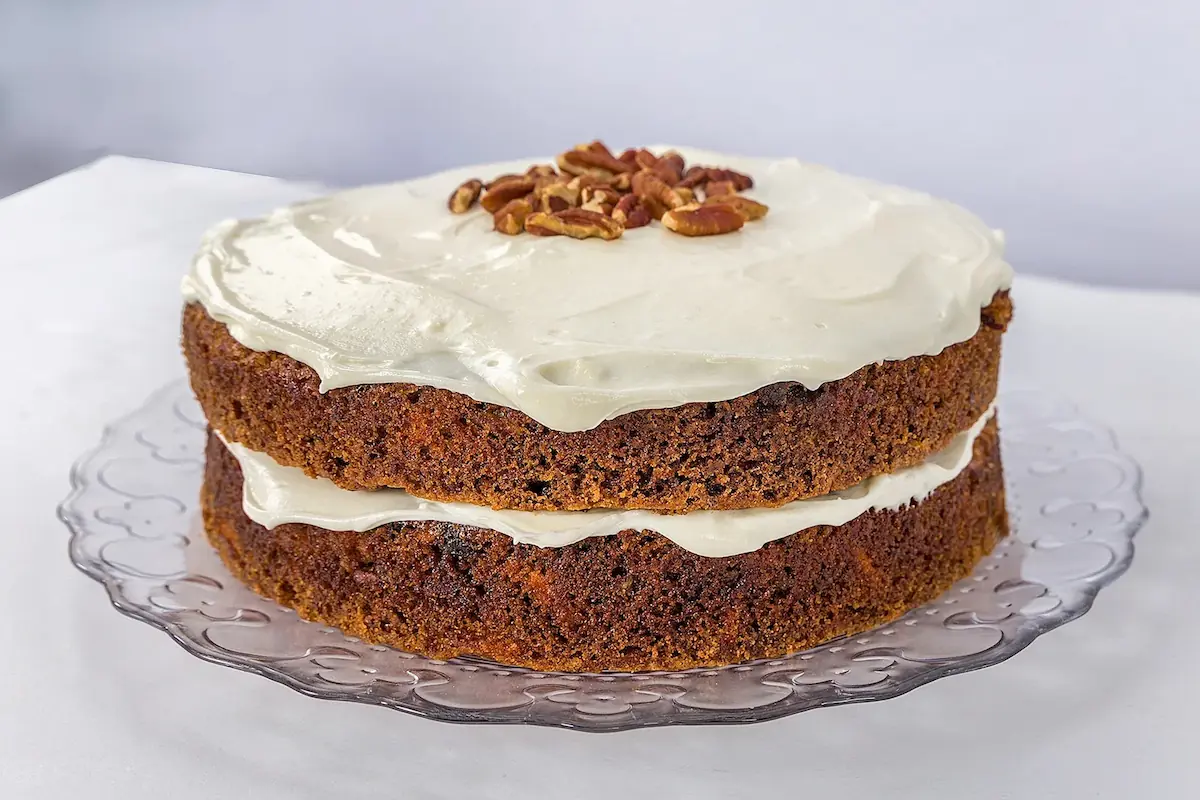 Moist two-layer carrot cake with thick cream cheese frosting and chopped pecans, displayed on a glass cake stand.